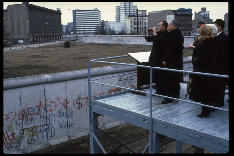 A group of men wearing black jackets stand at the edge of a platform and look toward gray buildings over a wall.